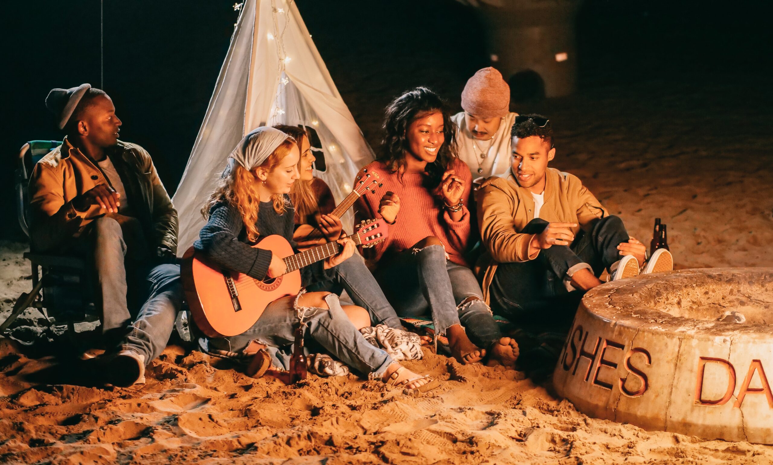 A group of friends sitting together on a beach at night, one playing guitar